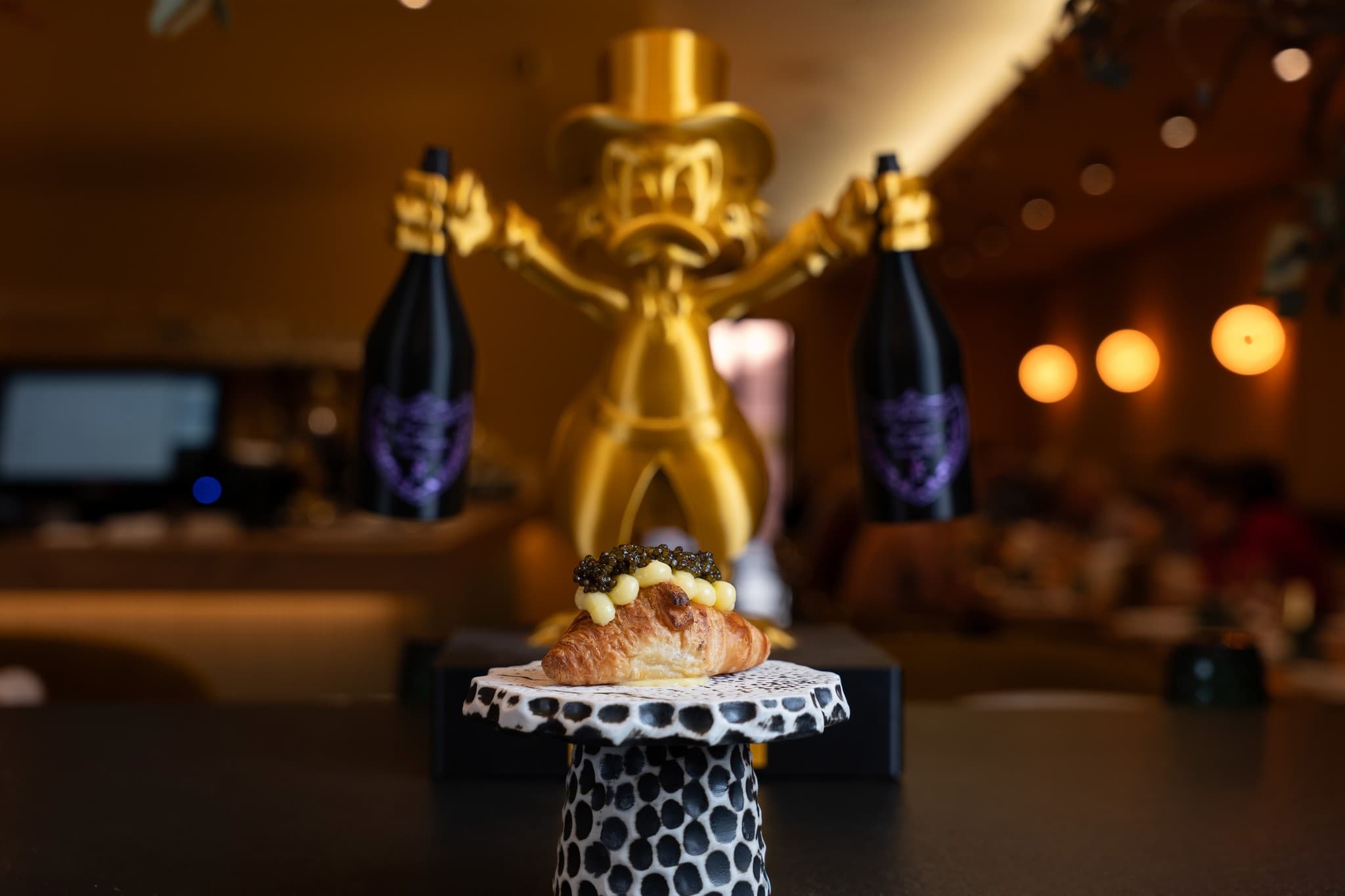 Croissant salado coronado con crema y caviar sobre un soporte de cerámica, con el comedor de Casa Varela desenfocado al fondo